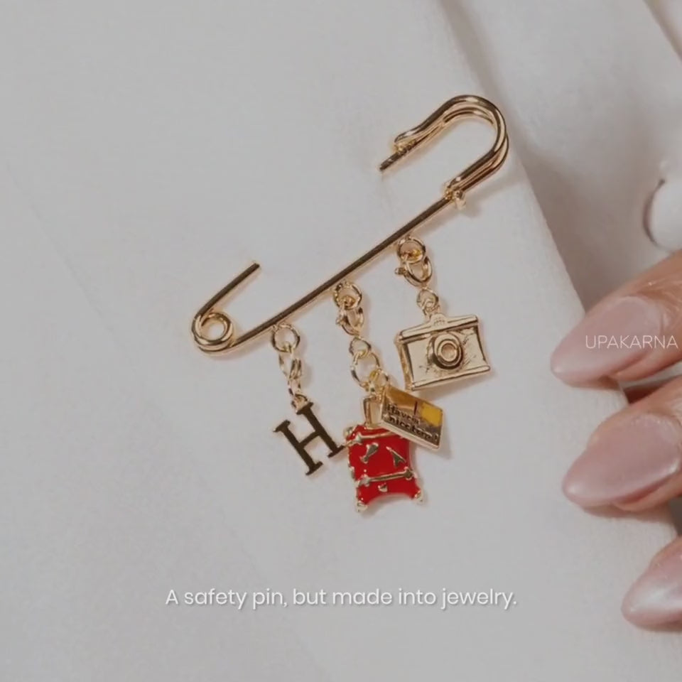 Video showcasing a gold-tone safety pin charm brooch with multiple dangling charms, including a letter, red dress, camera, and other decorative charms, shown as jewelry and styled on a handbag.
