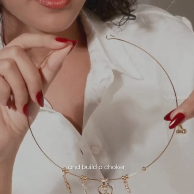 Close-up of a model in a white shirt holding a gold charm necklace wire with dangling heart and letter charms, demonstrating how it can be styled into a choker against a soft neutral background with the Upakarna logo.