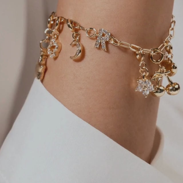 Close-up of a model wearing a gold charm bracelet with dangling moon, bow, flower, and teddy bear charms, styled on the wrist against a soft neutral background with the Upakarna logo.