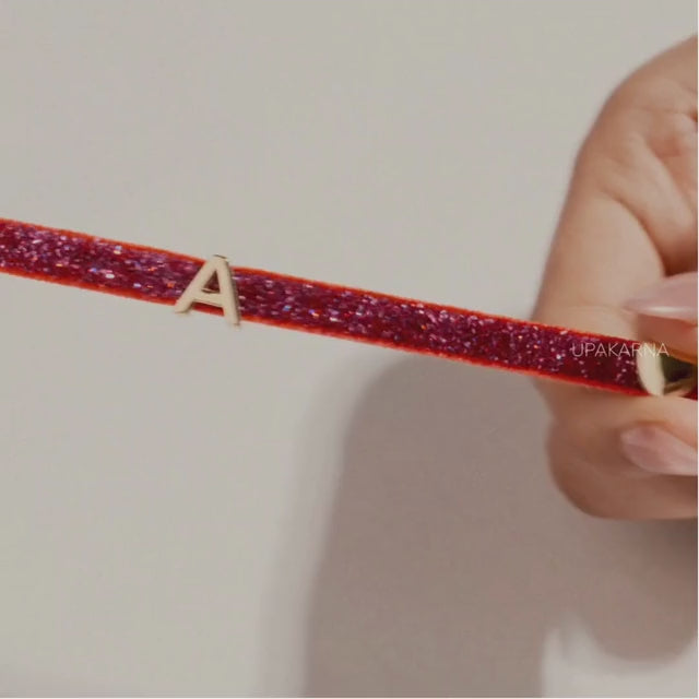 Close-up of hands sliding a gold letter A charm onto a red glitter shimmer tie-up bracelet, showcasing the personalized adjustable bracelet against a soft neutral background with the Upakarna logo.