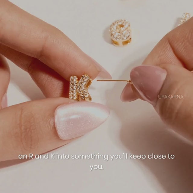A short promotional jewelry video featuring hands with light pink manicured nails threading sparkling crystal-encrusted gold letter charms onto a delicate chain. The video transitions to a close-up of a woman wearing the custom initial necklace against a white button-down shirt.