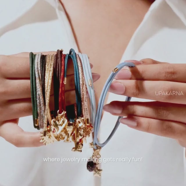 Close-up of hands sliding colorful gold-plated charms onto a light blue charm cord, featuring a flower, teacup, drink, and other playful pendants to showcase a customizable charm bracelet against a soft neutral background with the Upakarna logo.
