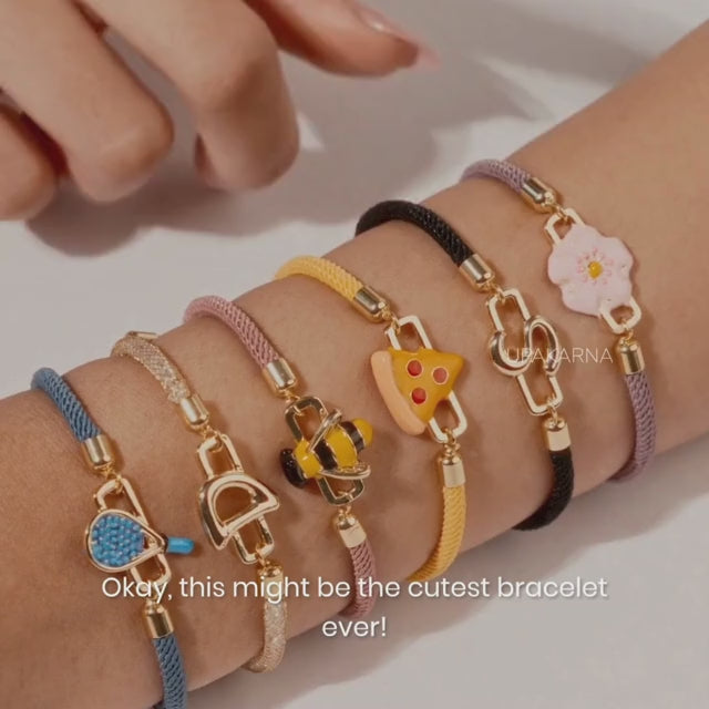 Close-up of a model in a white shirt adjusting a layered gold charm bracelet with heart and letter pendants, showcasing an elegant everyday styling look against a soft neutral background with the Upakarna logo.