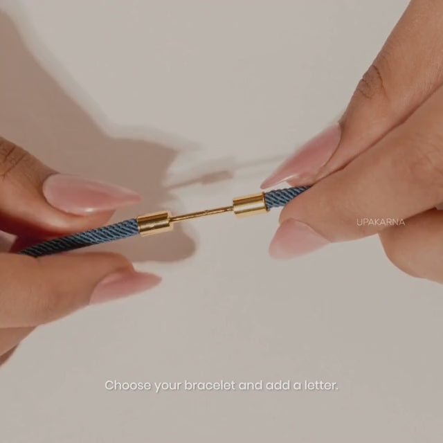 Close-up of hands connecting a navy braided bracelet with a gold letter charm clasp, demonstrating the customizable bracelet design against a warm neutral background with the text.
