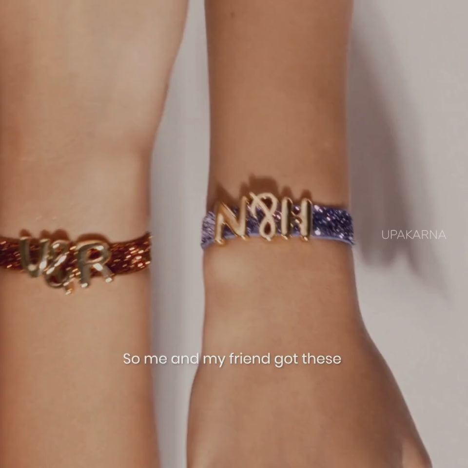 Video showing a blue shimmery band being personalized with gold-tone initial charms and colorful decorative charms, including a pink flower and heart, before the finished charm band is worn on the wrist.
