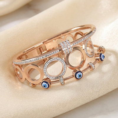 Rose gold anti-tarnish bangle stack featuring crystal-studded geometric bangles and blue evil eye accents, elegantly displayed on a soft satin fabric background.