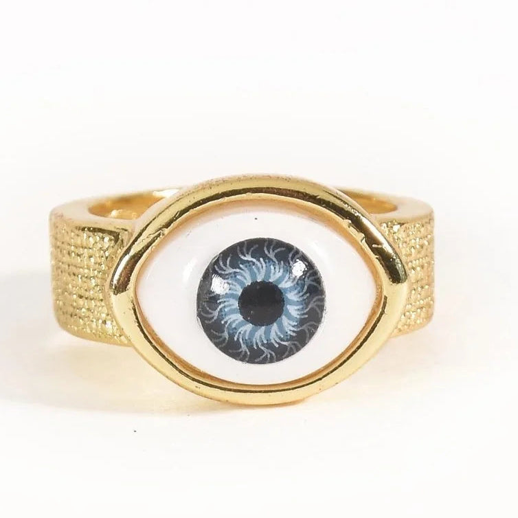 The image shows a gold-toned statement ring featuring a large blue-and-black evil-eye motif at the center, framed by a smooth gold oval. The wide textured gold band has fine dotted detailing along its surface. It is photographed against a clean white background.
