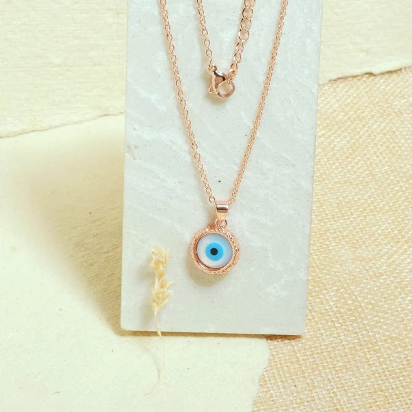The image shows a rose-gold evil-eye necklace displayed on a small white jewelry card. It features a delicate chain and a round pendant with a blue-and-white evil-eye motif, surrounded by a rose-gold textured border. The card is placed on a soft cream fabric background with a warm, minimal aesthetic.