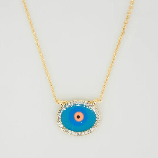 The image shows a delicate gold-toned necklace with a fine chain and an oval evil-eye pendant. The pendant has a vivid turquoise-blue center, a peach and black eye motif, and a sparkling clear-stone border. It is photographed against a clean light-gray/white background.
