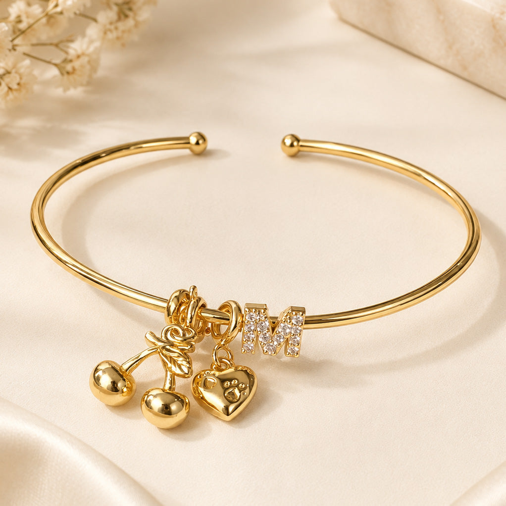 Gold open cuff bangle featuring a sparkling letter M charm with cherry and heart charms, styled on a cream satin surface.